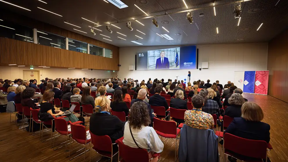 European Commission, Commissioner Mr. Olivér Várhelyi on big screen. Participants are seated in an auditorium.